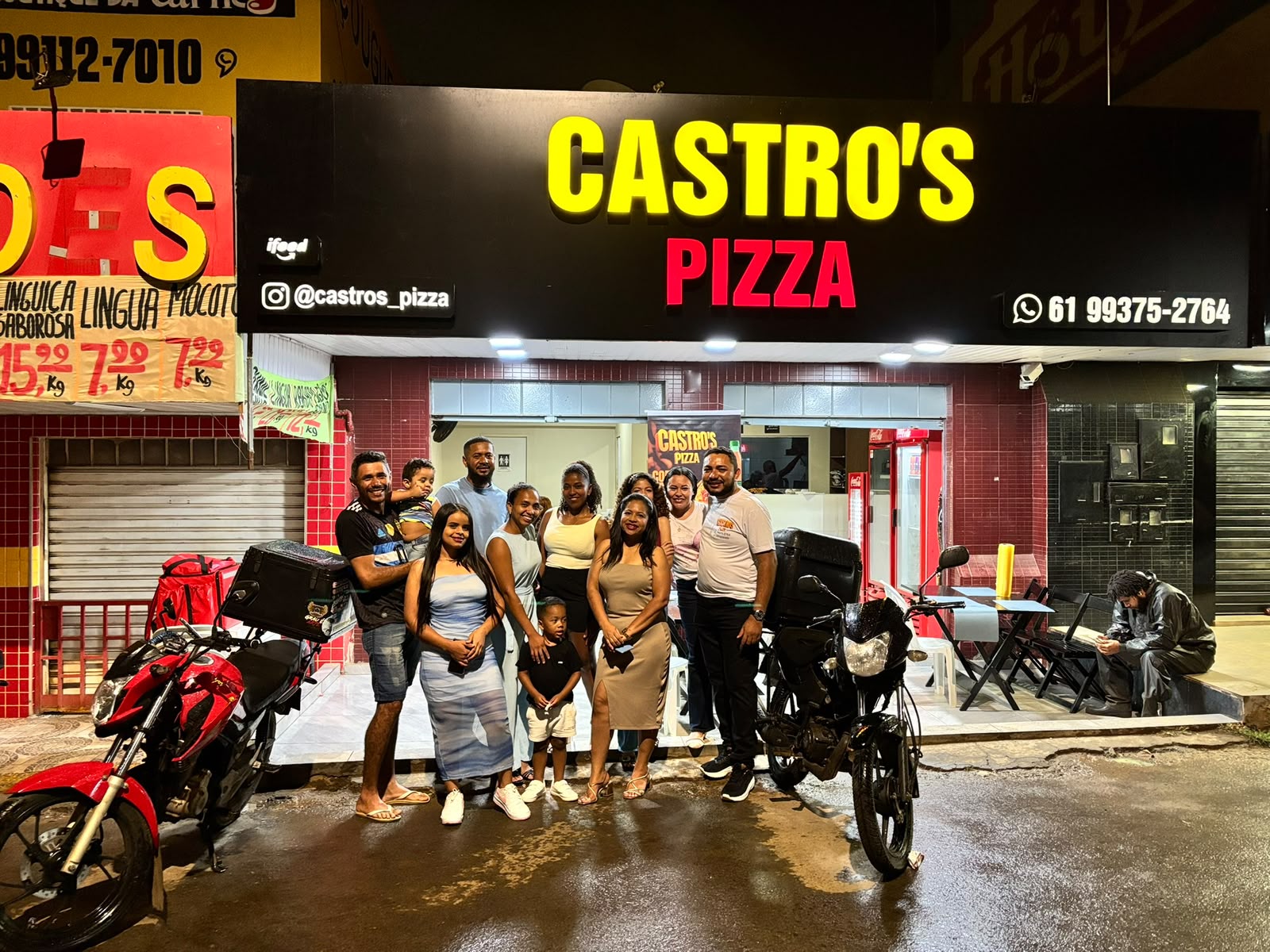 Fachada iluminada da Castro's Pizza à noite, com pessoas reunidas na entrada e motos de entrega estacionadas